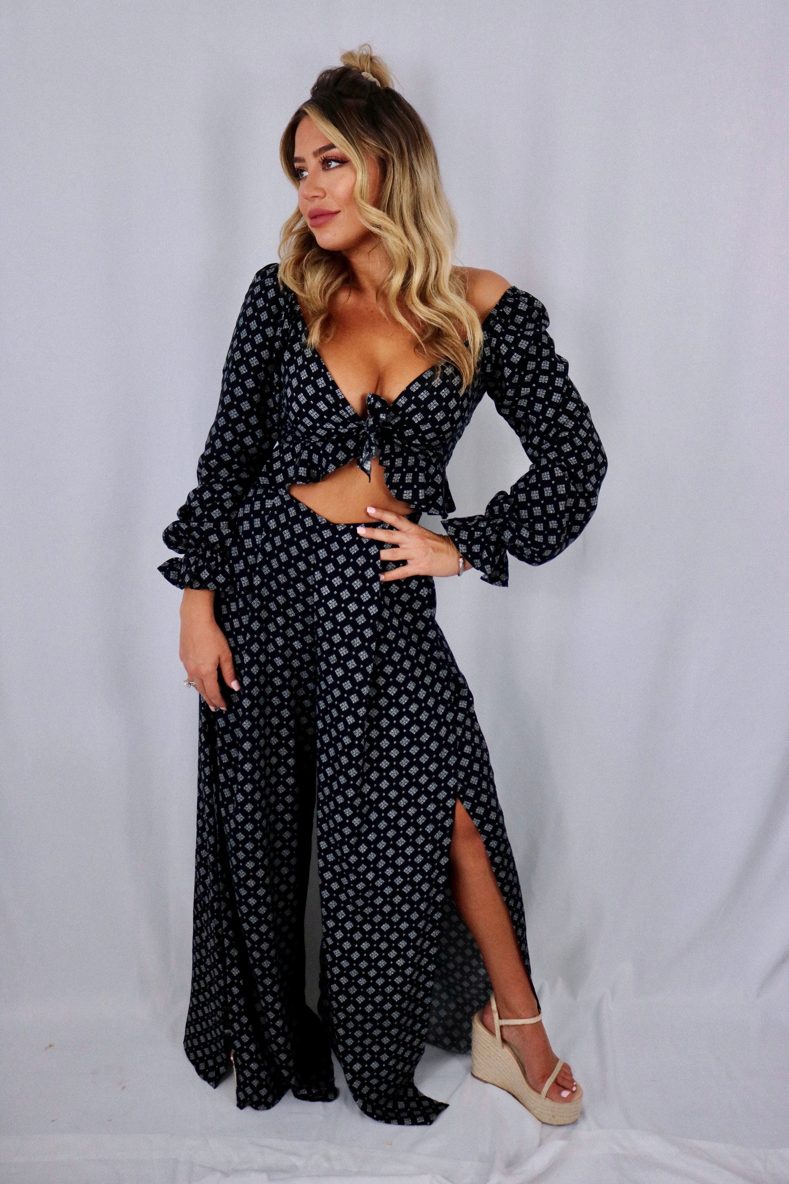 Blonde girl models a black, flowy matching set for Scarlette The Label, an online fashion boutique for women. The matching set includes a black crop tie top and black flare, flowy pants. Each sold separately.