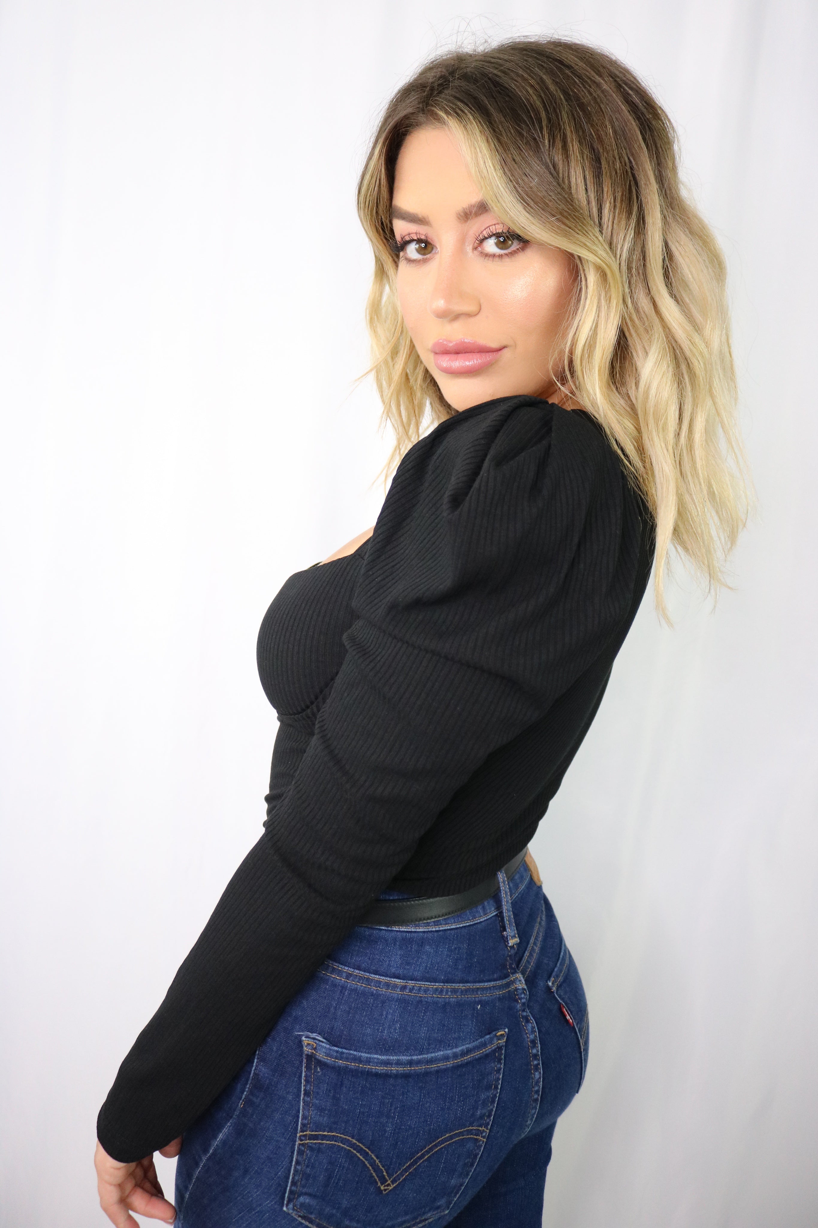 Blonde girl models a long sleeve bodysuit in black for Scarlette The Label, an online fashion boutique for women. The black bodysuit has puff sleeves. Paired with dark denim jeans.