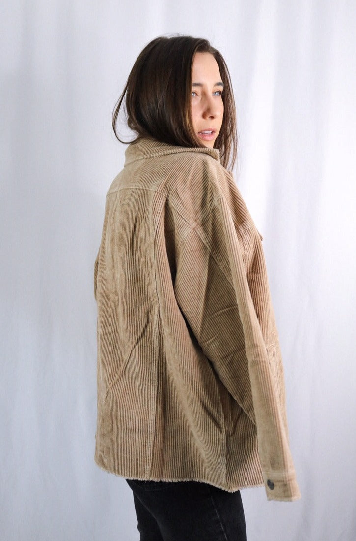 Brunette girl models buttoned corduroy jacket in the color tan for Scarlette The Label, an online fashion boutique for women. The corduroy jacket is an oversized street style jacket. Paired with a black crop top, dark denim jeans.
