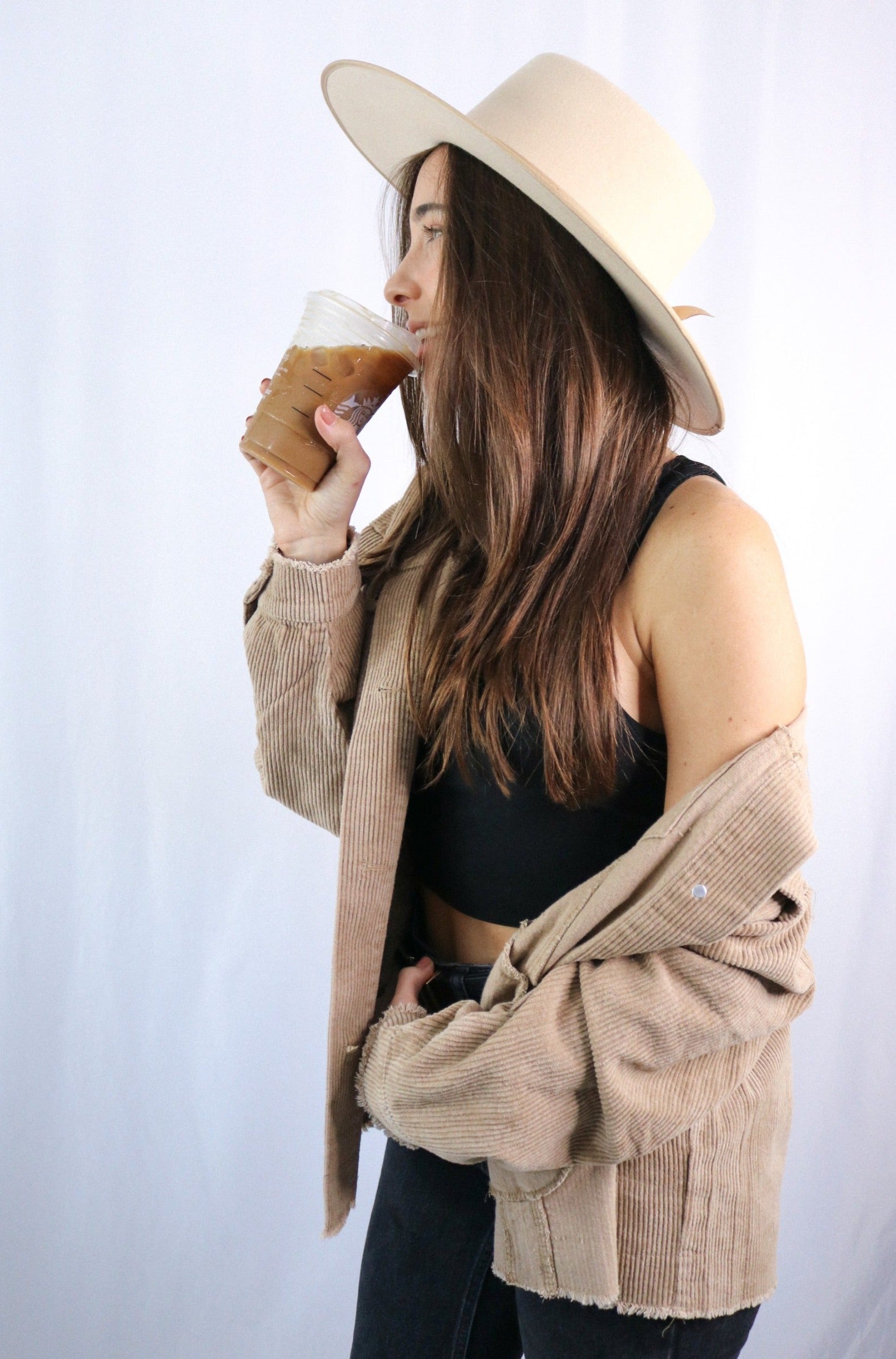 Brunette girl models buttoned corduroy jacket in the color tan for Scarlette The Label, an online fashion boutique for women. The corduroy jacket is an oversized street style jacket. Paired with a black crop top, dark denim jeans, and a wide-brimmed rancher hat.