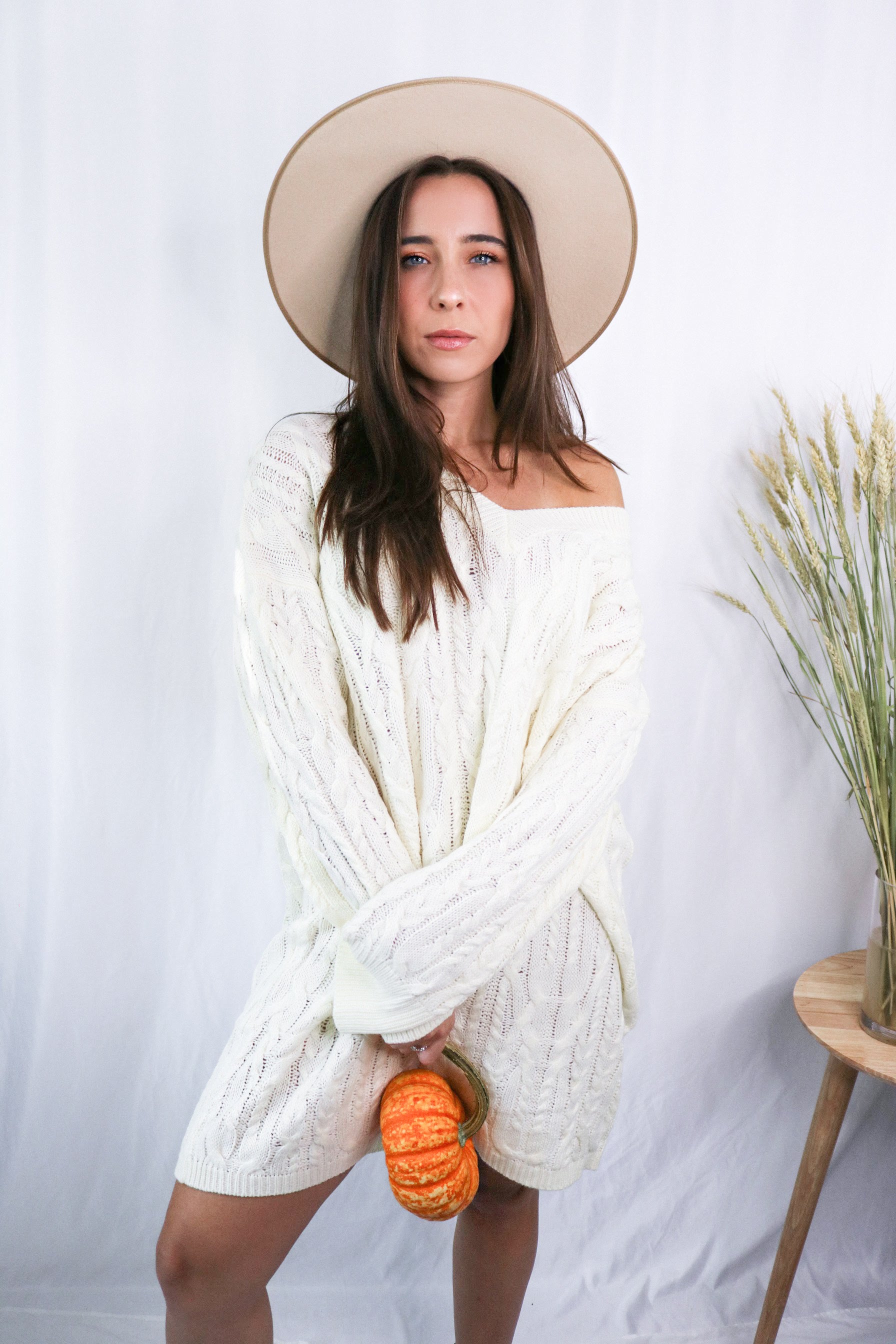 Brunette girl models a mid-length cable knit sweater dress in the color ivory for Scarlette The Label, an online fashion boutique for women. Paired with a beige wide-brimmed rancher hat.