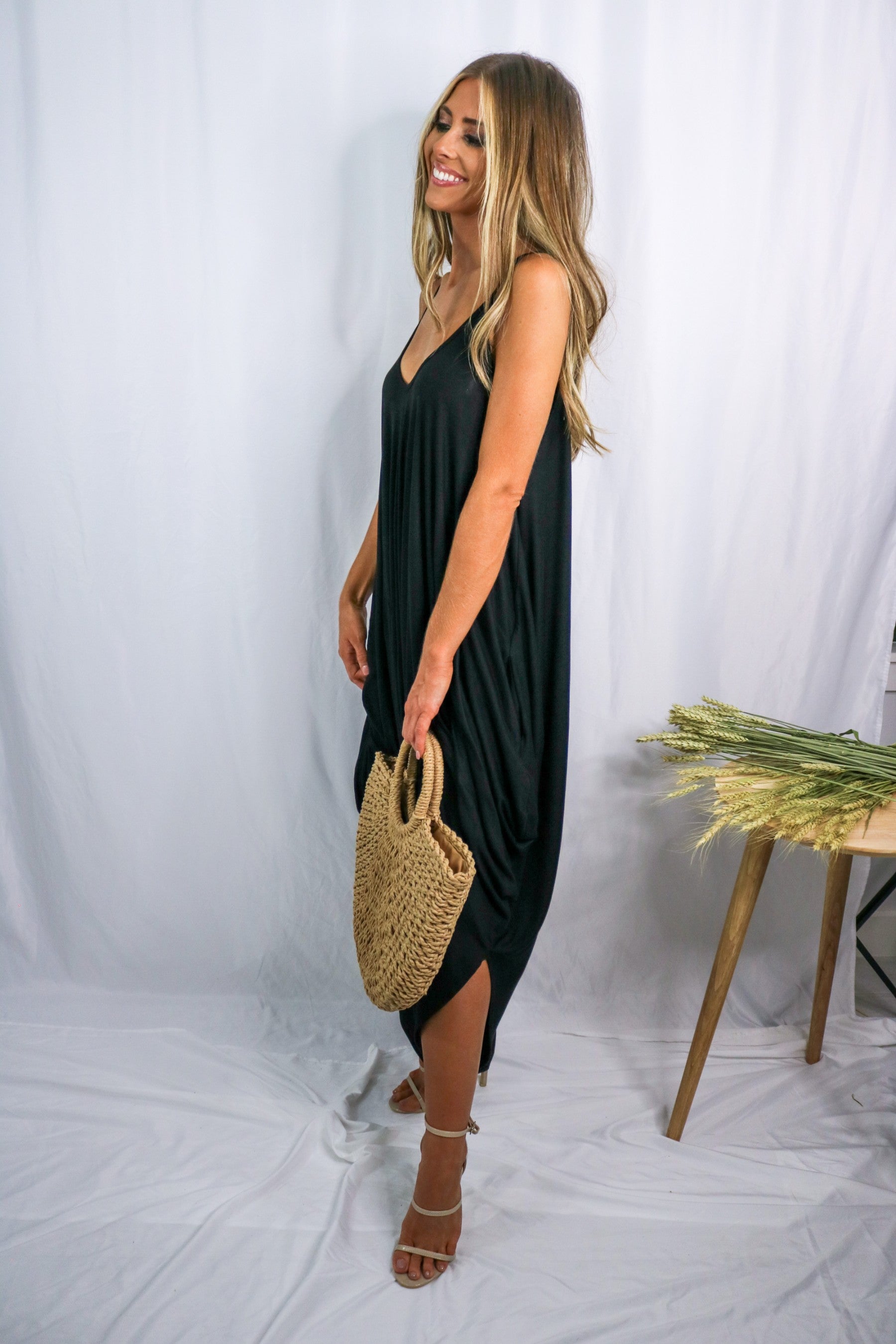Blonde girl modeling a black boxy hi lo jumpsuit for Scarlette The Label, an online fashion boutique for women. Black jumpsuit has pockets and is lightweight and flowy. Paired with large straw bag.
