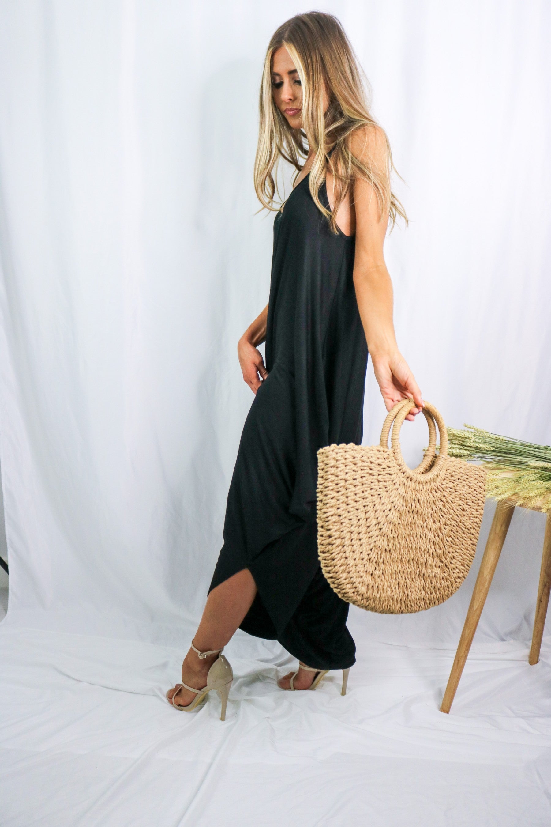 Blonde girl modeling a black boxy hi lo jumpsuit for Scarlette The Label, an online fashion boutique for women. Black jumpsuit has pockets and is lightweight and flowy. Paired with large straw bag.