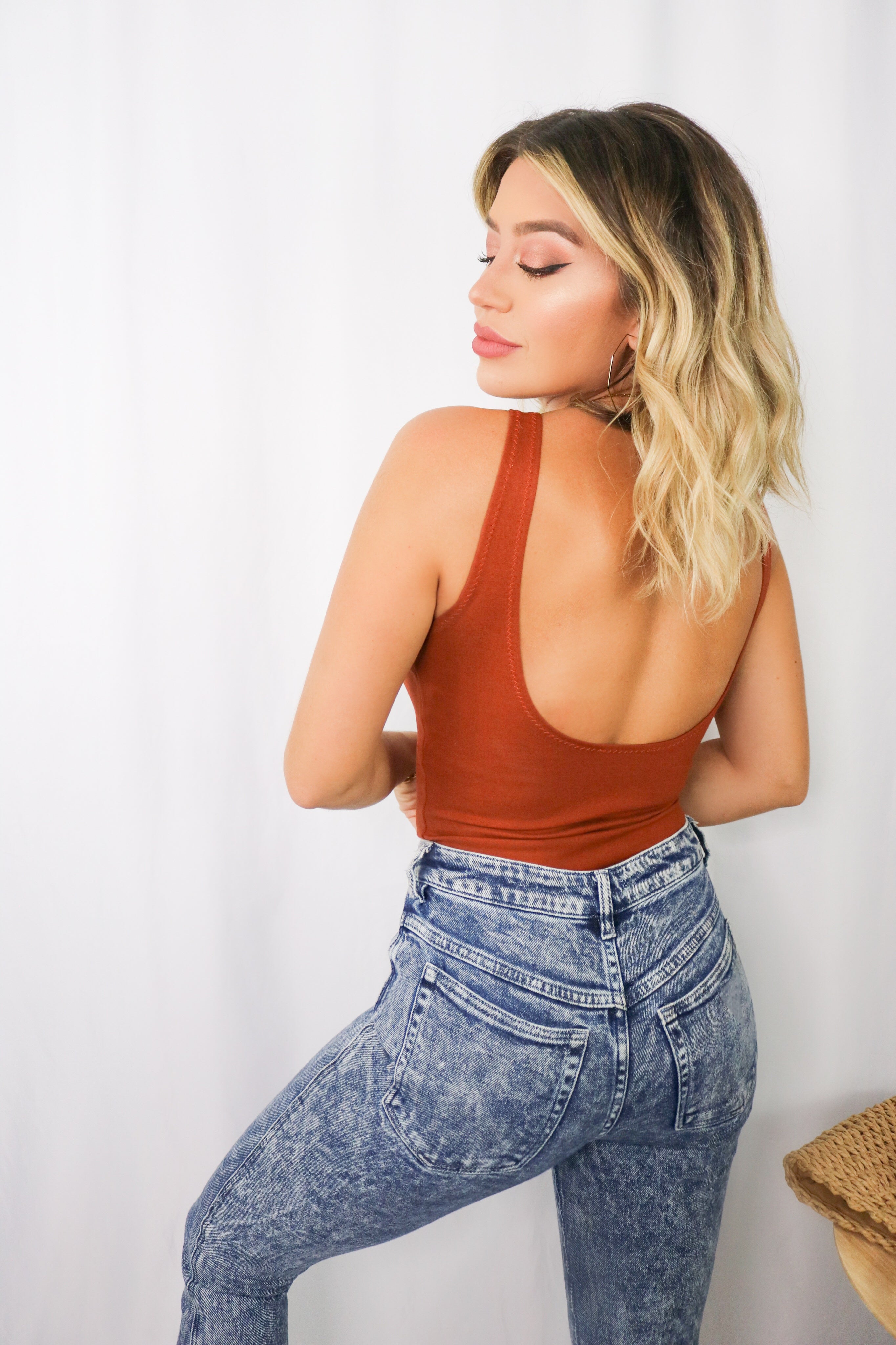 Blonde girl models a plain ponte bodysuit in the color rust for Scarlette The Label, an online fashion boutique for women. Paired with dark denim jeans and a beige wide-brimmed rancher hat.