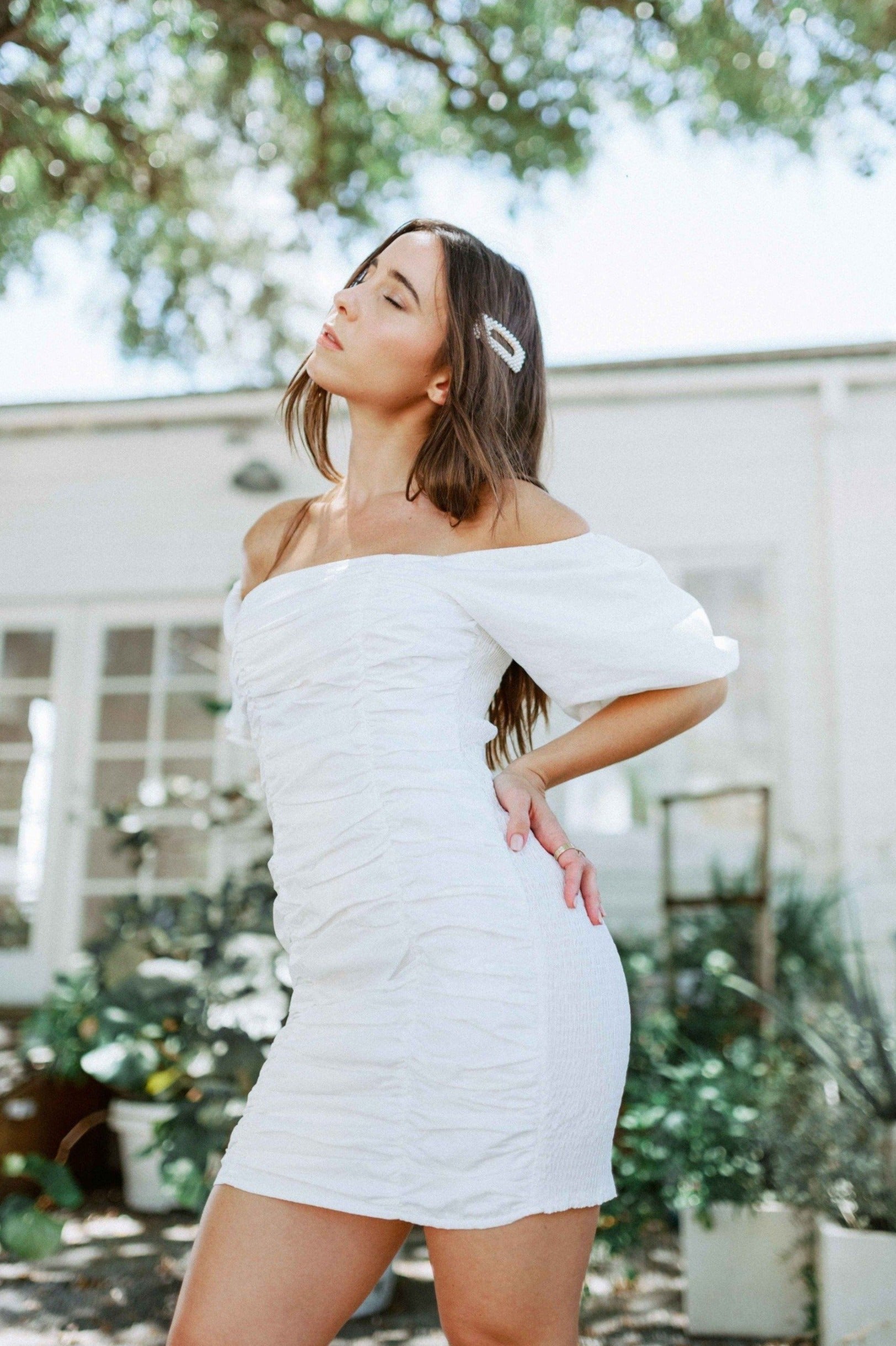 Brunette girl models white ruched mini dress with puff sleeves for Scarlette The Label, an online fashion boutique for women. White mini dress can be worn off the shoulders or regular.