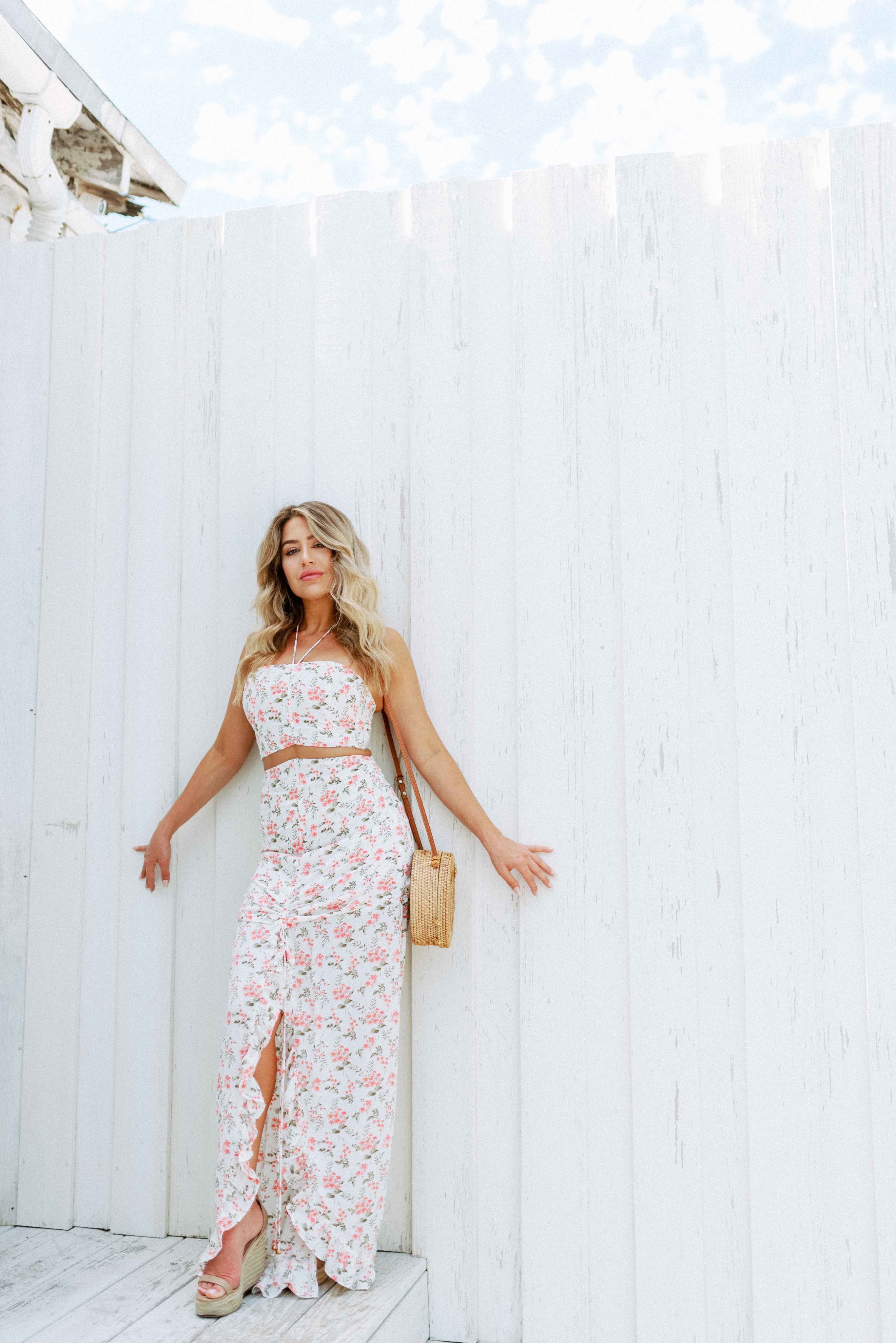 Blonde girl models a floral maxi skirt matching set for Scarlette The Label, an online fashion boutique for women. The matching skirt set has a halter crop top and a long slitted maxi skirt. Sold separately. Paired with straw purse.