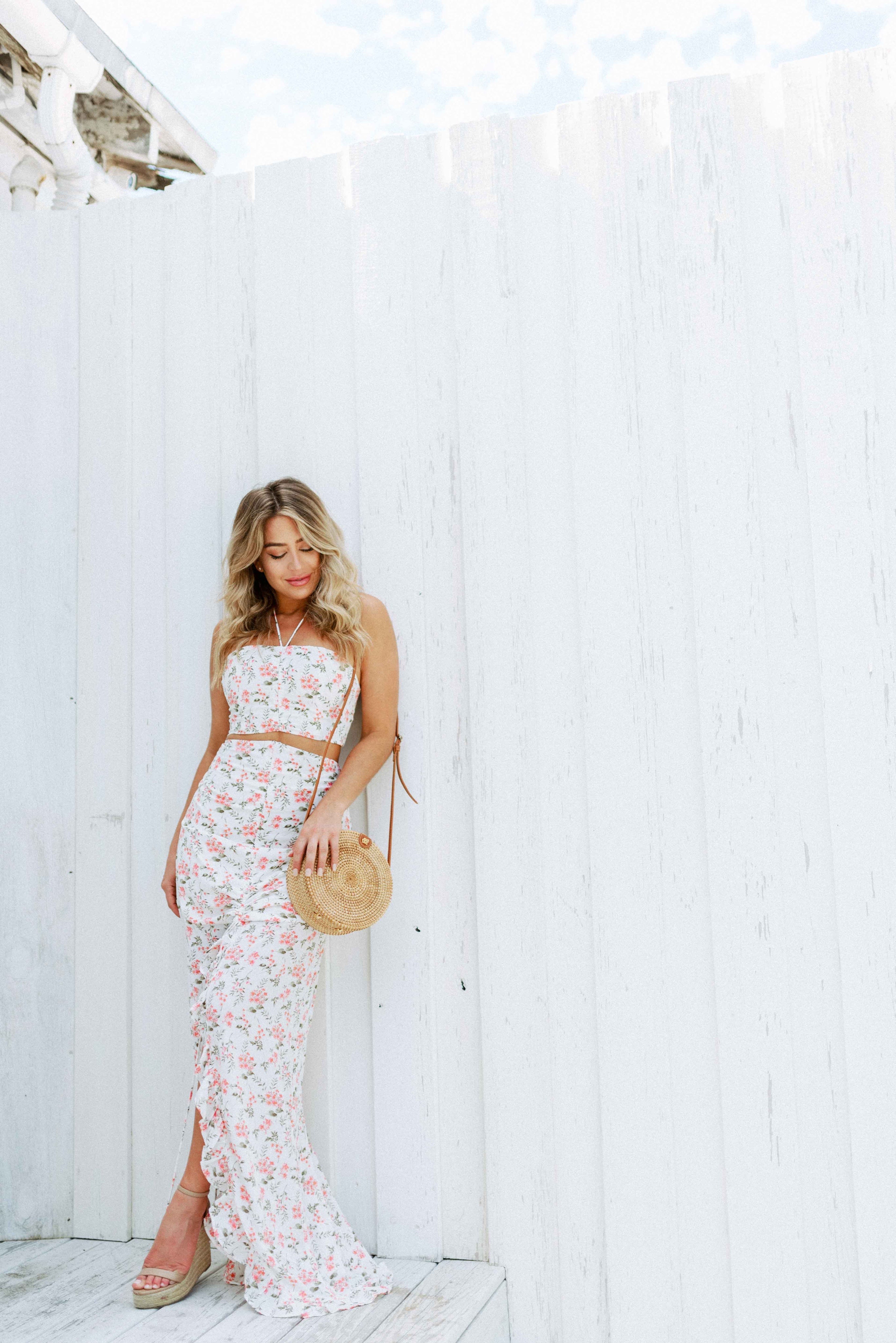 Blonde girl models a floral maxi skirt matching set for Scarlette The Label, an online fashion boutique for women. The matching skirt set has a halter crop top and a long slitted maxi skirt. Sold separately. Paired with straw purse.