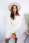 Brunette girl models a mid-length cable knit sweater dress in the color ivory for Scarlette The Label, an online fashion boutique for women. Paired with a beige wide-brimmed rancher hat.
