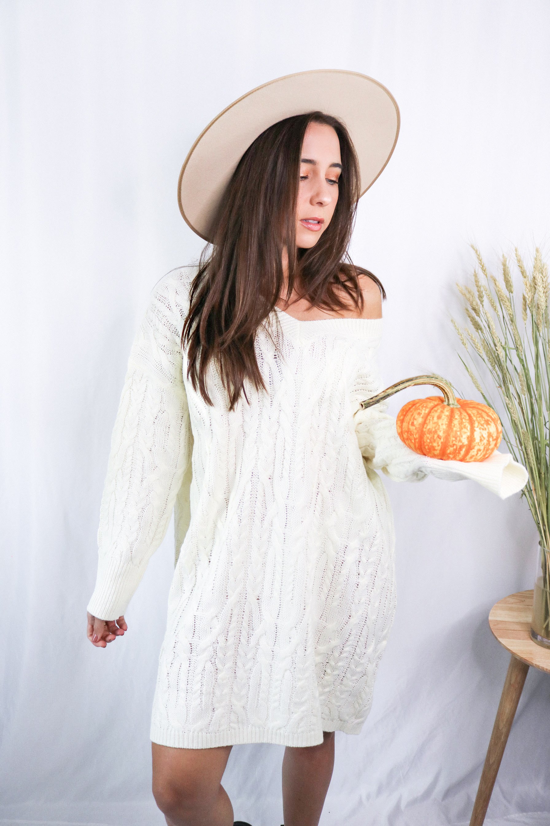 Brunette girl models a mid-length cable knit sweater dress in the color ivory for Scarlette The Label, an online fashion boutique for women. Paired with a beige wide-brimmed rancher hat.
