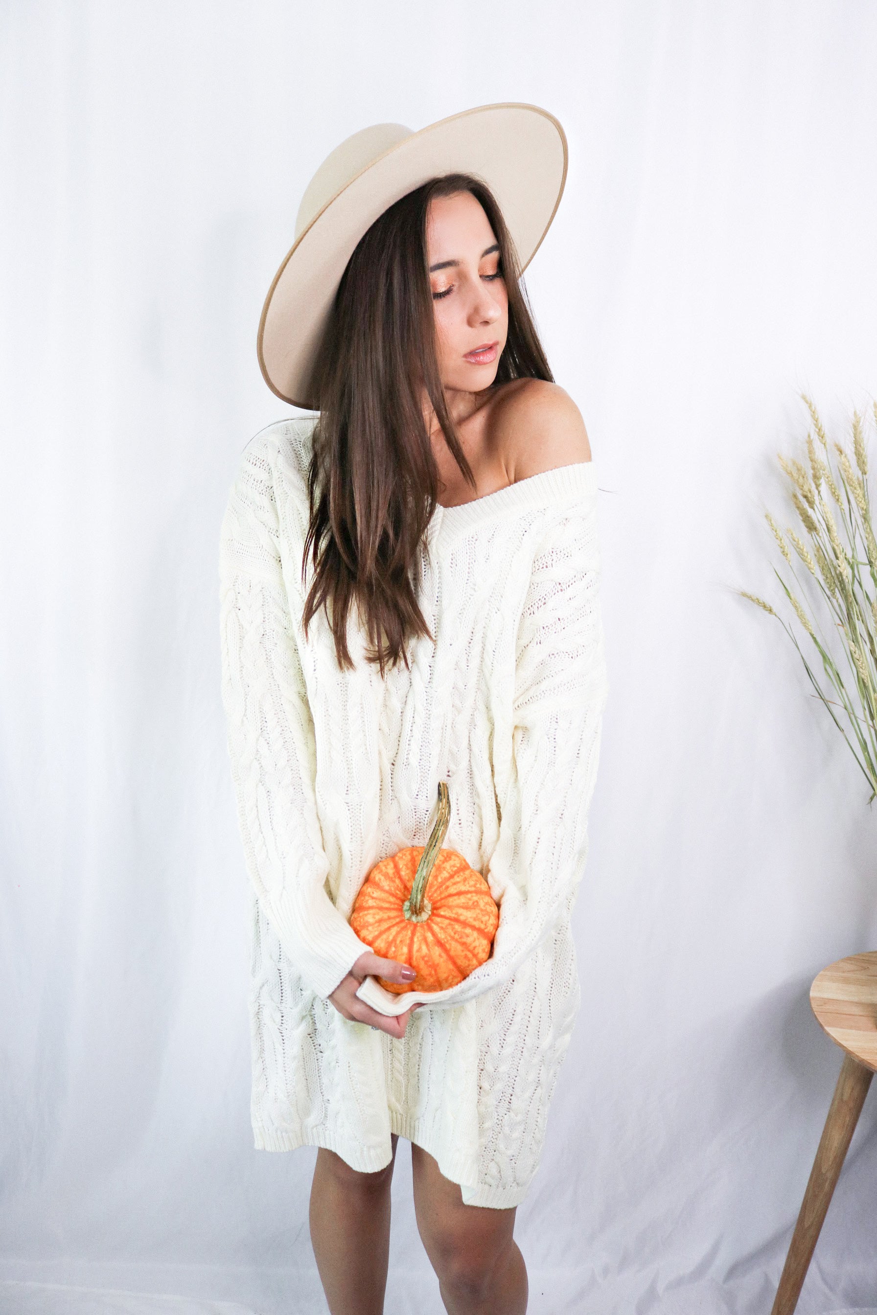 Brunette girl models a mid-length cable knit sweater dress in the color ivory for Scarlette The Label, an online fashion boutique for women. Paired with a beige wide-brimmed rancher hat.