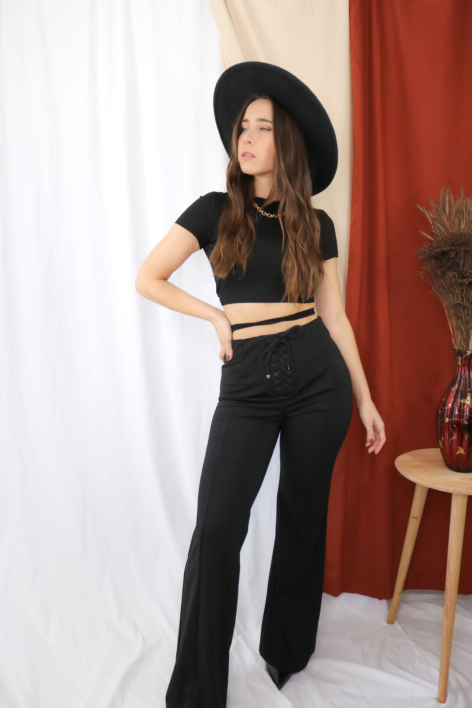 Brunette models outfit for Scarlette the Label, an online fashion boutique for women. Wears an open back black crop top and flare wide leg tie pants in black. Paired with wide brim rancher hat.