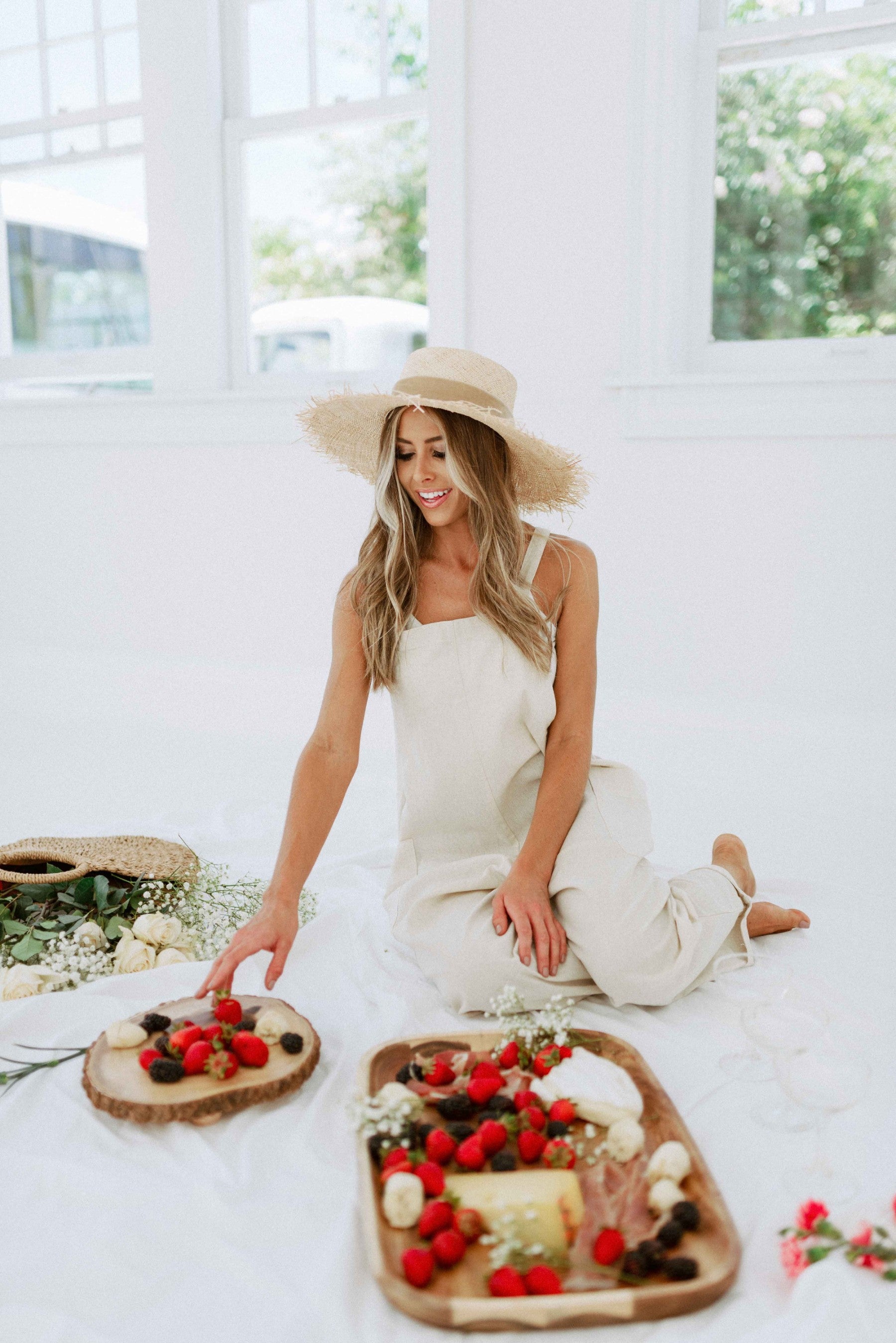 Blonde girl modeling pocketed overalls in color oatmeal for Scarlette The Label, an online fashion boutique for women. The overalls are light beige and have open sides. Paired with a wide-brimmed straw hat.