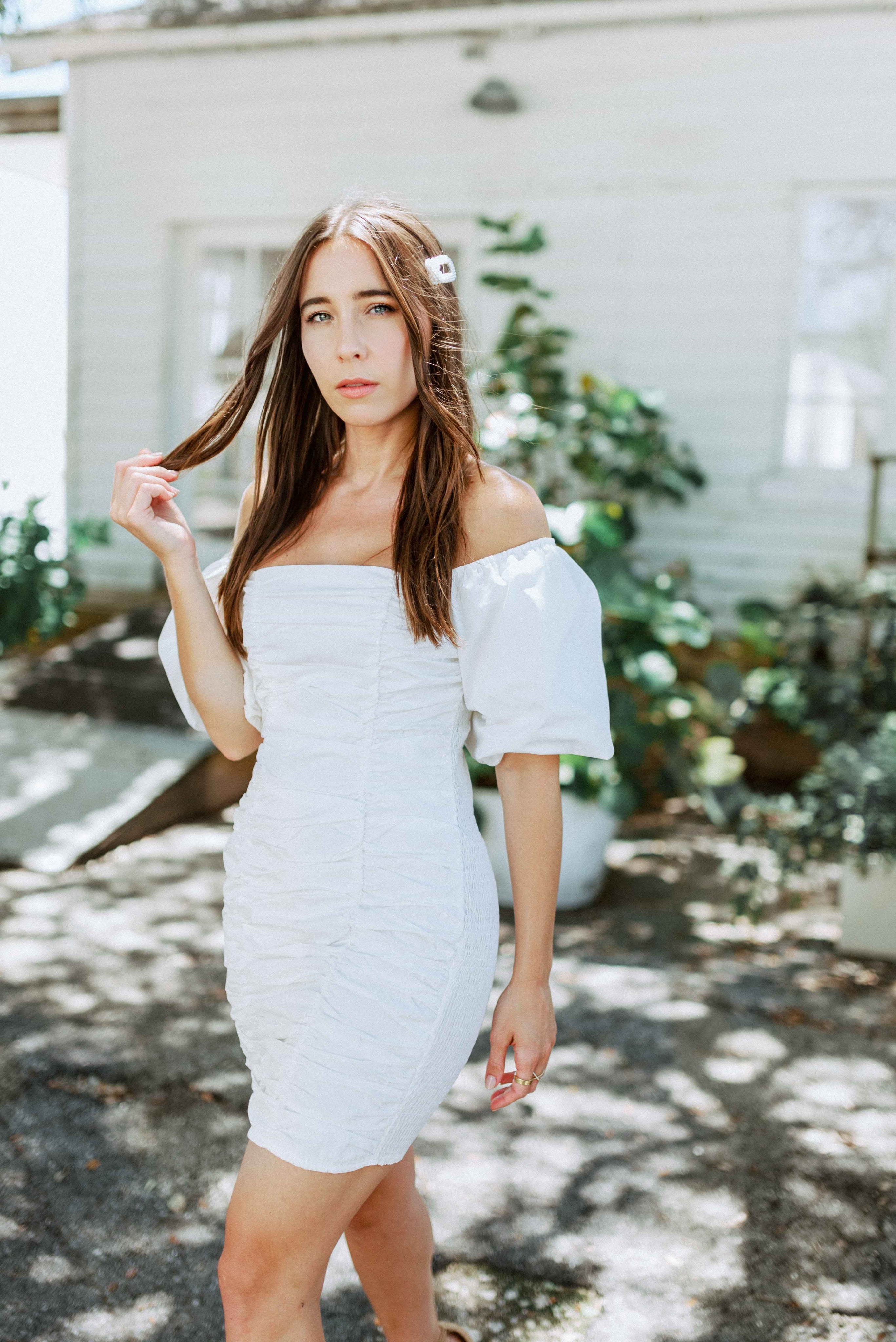 Brunette girl models white ruched mini dress with puff sleeves for Scarlette The Label, an online fashion boutique for women. White mini dress can be worn off the shoulders or regular.