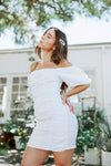 Brunette girl models white ruched mini dress with puff sleeves for Scarlette The Label, an online fashion boutique for women. White mini dress can be worn off the shoulders or regular.