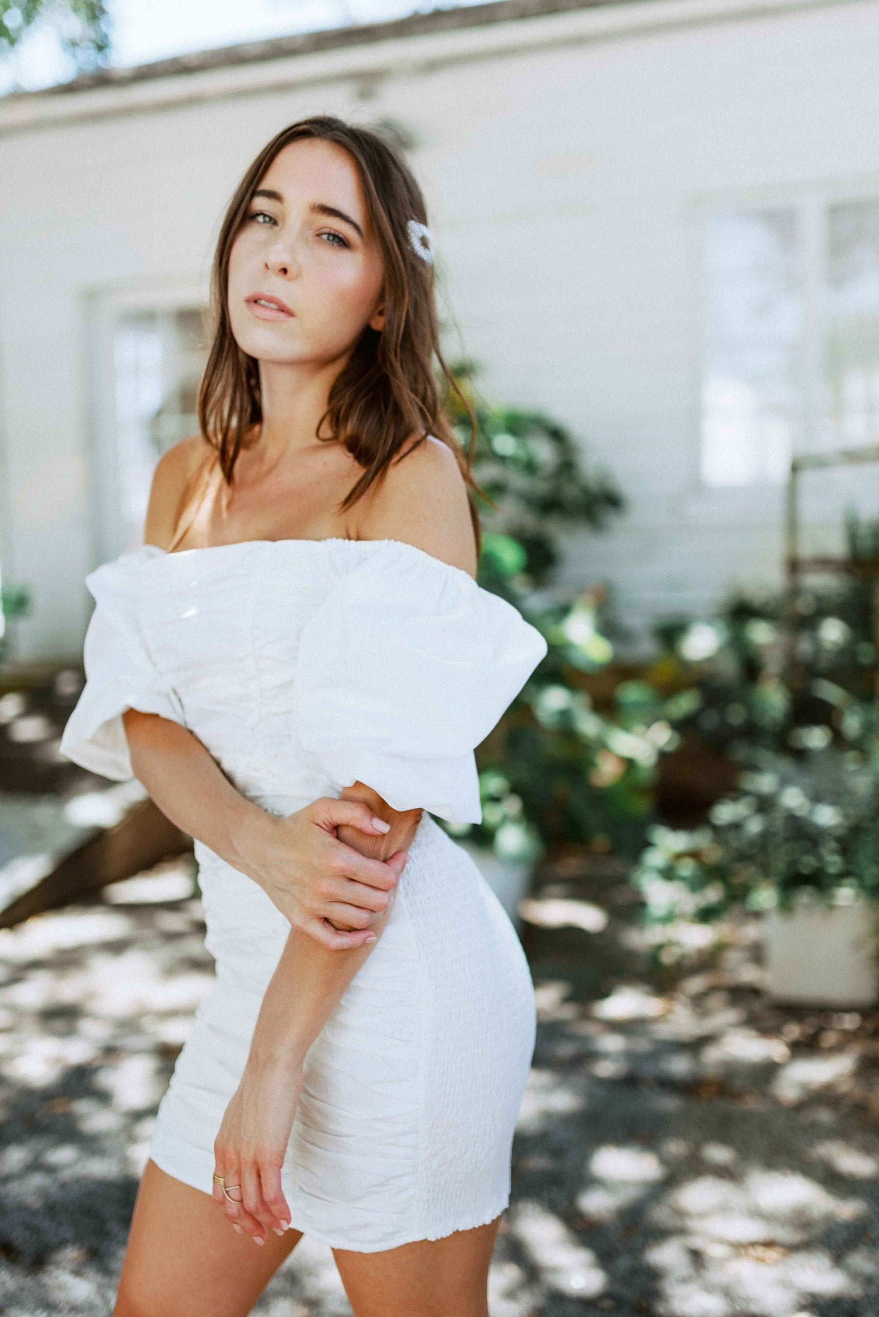 Brunette girl models white ruched mini dress with puff sleeves for Scarlette The Label, an online fashion boutique for women. White mini dress can be worn off the shoulders or regular.